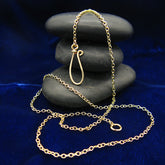 A gold-plated chain necklace with a handmade loop clasp, draped over a stack of dark gray stones on a deep blue velvet fabric background.