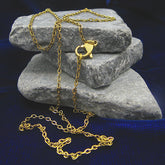 A gold flat cable chain necklace with a lobster clasp, casually draped over three stacked gray stones against a dark blue textured background.