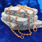 A copper figure 8 chain necklace with a lobster clasp, draped over a stack of gray stones on a textured blue fabric background.