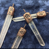 Four clear quartz crystal point pendants with ornate copper metal caps featuring symbolic engravings, arranged on a textured navy blue fabric background.