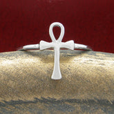 Front view of a silver ring in the shape of an Egyptian ankh symbol resting on a rounded textured golden ceramic prop with a dark red background.