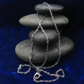 A silver stainless steel chain necklace with lobster clasp draped over a stack of smooth dark gray stones on a deep blue fabric background.