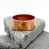 Gently hammered pure copper band ring resting on stacked grey stones with white background.