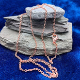 A delicate copper chain necklace draped over stacked gray stones on a textured blue fabric background.