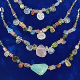 A set of three layered beaded necklaces displayed on a textured royal blue velvet background. The necklaces feature a variety of colorful raw gemstones, including a prominent light pink stone, a large light green stone, a pinkish-purple stone, and silver charms including a pentagram and an eye of providence.