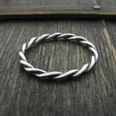 Overhead view of a slender silver ring with a braided rope pattern design sitting on the top edge of a dark greenish weathered wooden surface.