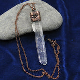 Necklace with pendant of clear quartz point with handmade copper cap with evil eye design and copper chain draped over a smooth grey stone on a dark blue cloth background.