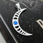 Sterling silver pendant in shape of crescent moon with blue cabochon and a cut-out moon-phases design on a weathered black wooden surface with white graphics.