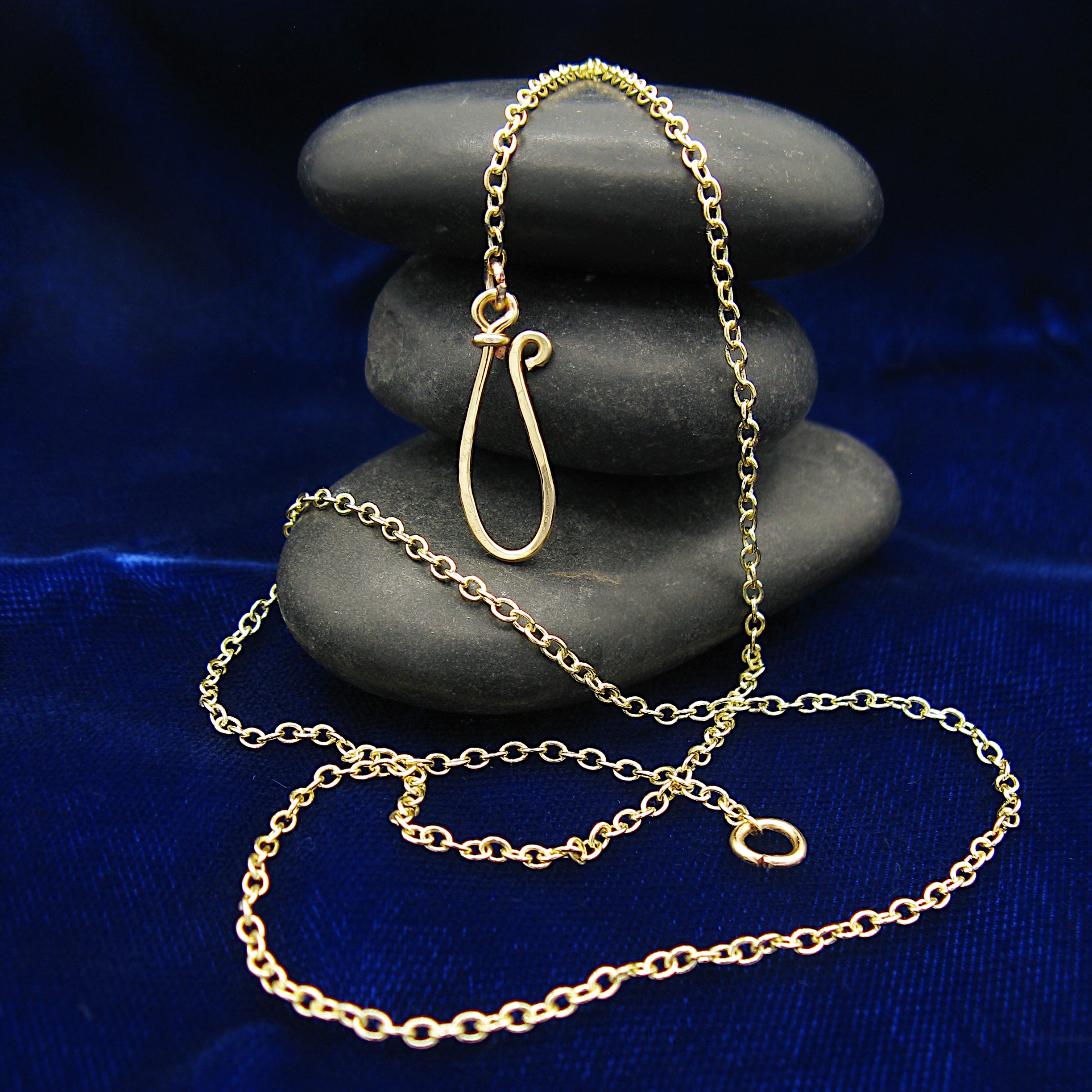 A gold-plated chain necklace with a handmade loop clasp, draped over a stack of dark gray stones on a deep blue velvet fabric background.