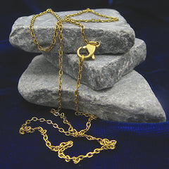 A gold flat cable chain necklace with a lobster clasp, casually draped over three stacked gray stones against a dark blue textured background.
