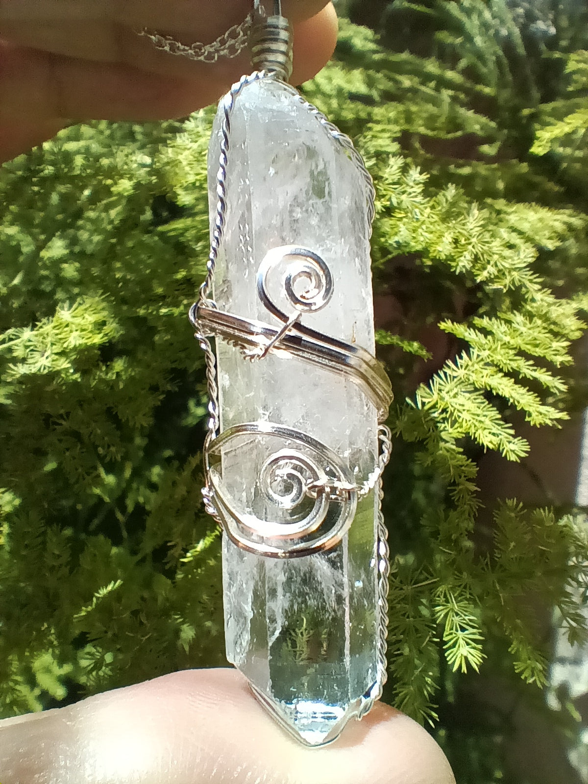 A clear quartz crystal pendant with silver wire swirl accents, held in a hand against a background of green foliage.