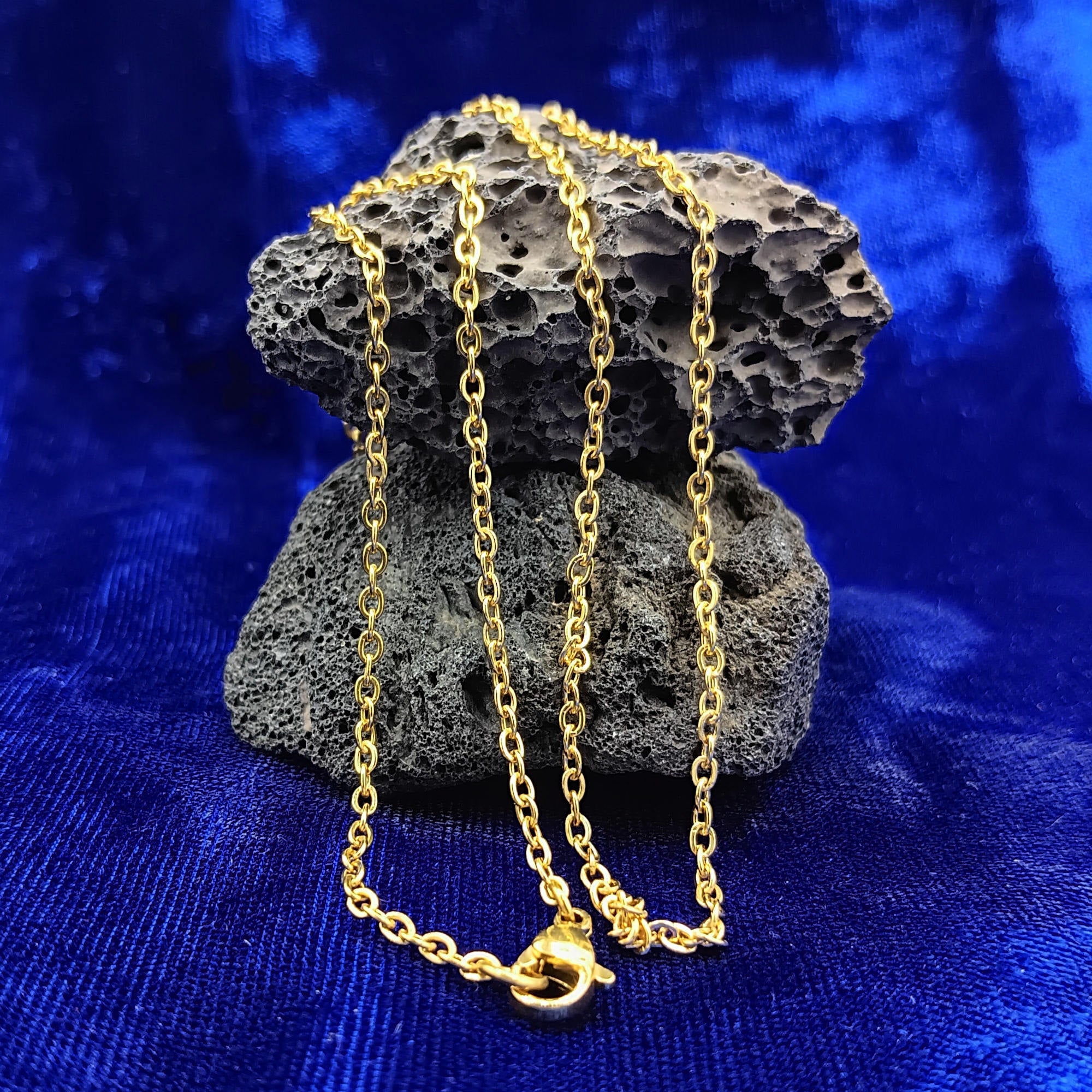 A gold chain necklace draped over a dark porous rock, set against a vibrant blue velvet fabric background.