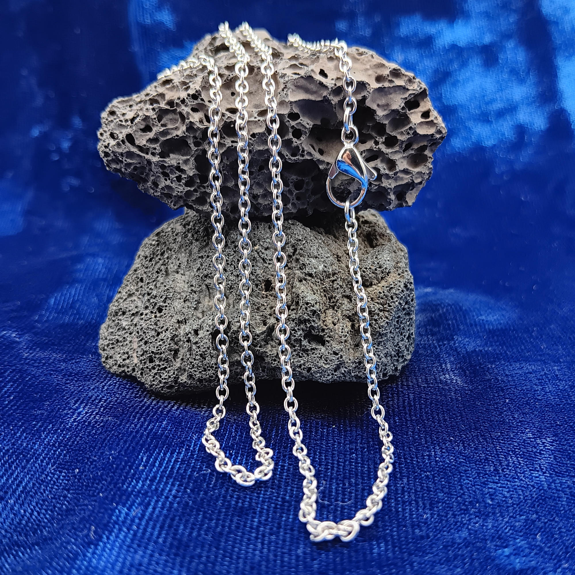 A stainless steel chain necklace draped over a dark porous rock, set against a rich blue velvet fabric background.