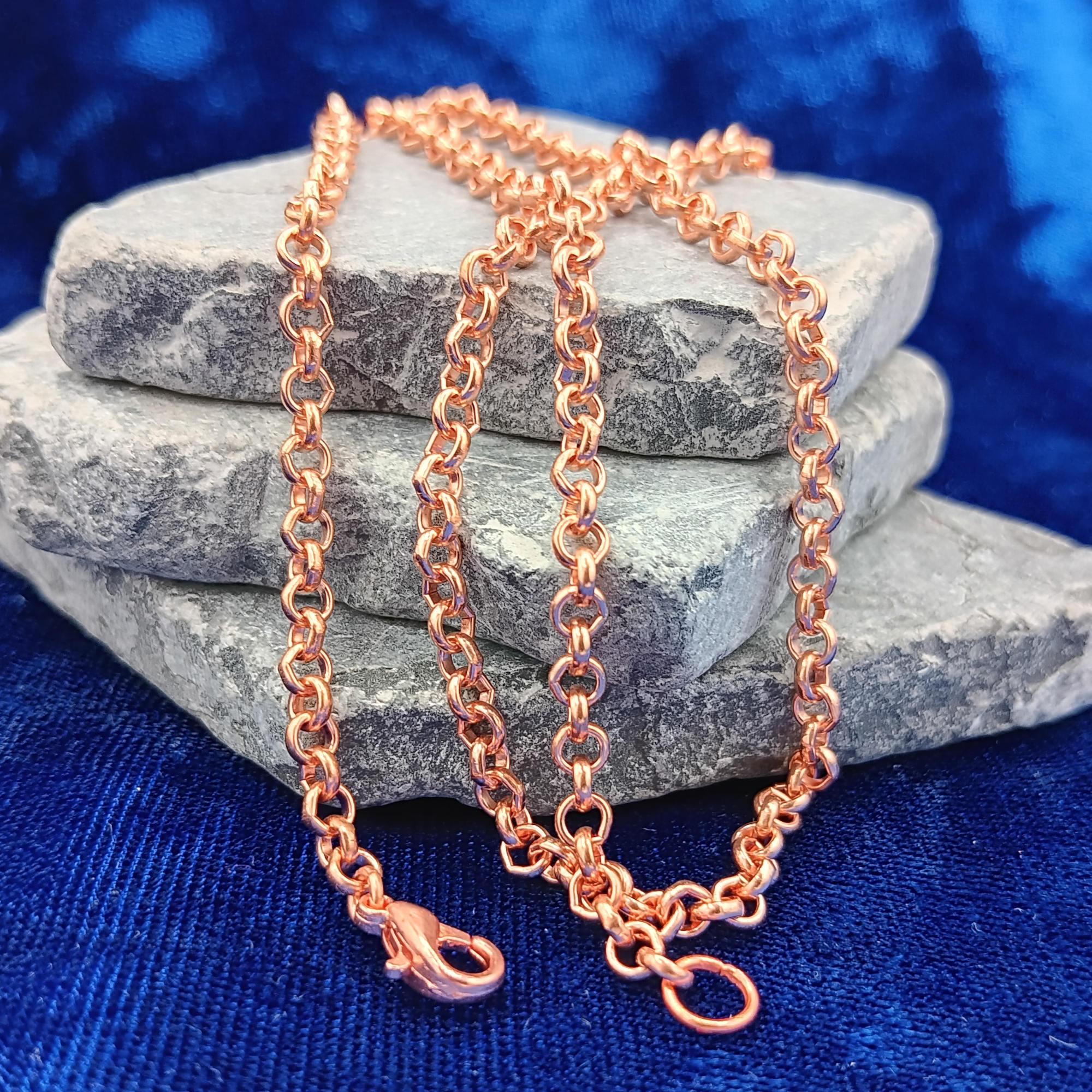A copper rolo chain necklace draped over stacked gray stones on a textured blue fabric background.