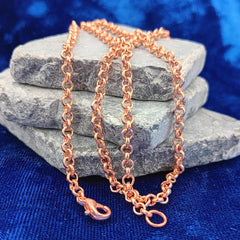 A copper rolo chain necklace draped over stacked gray stones on a textured blue fabric background.