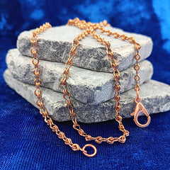 A copper figure 8 chain necklace with a lobster clasp, draped over a stack of gray stones on a textured blue fabric background.