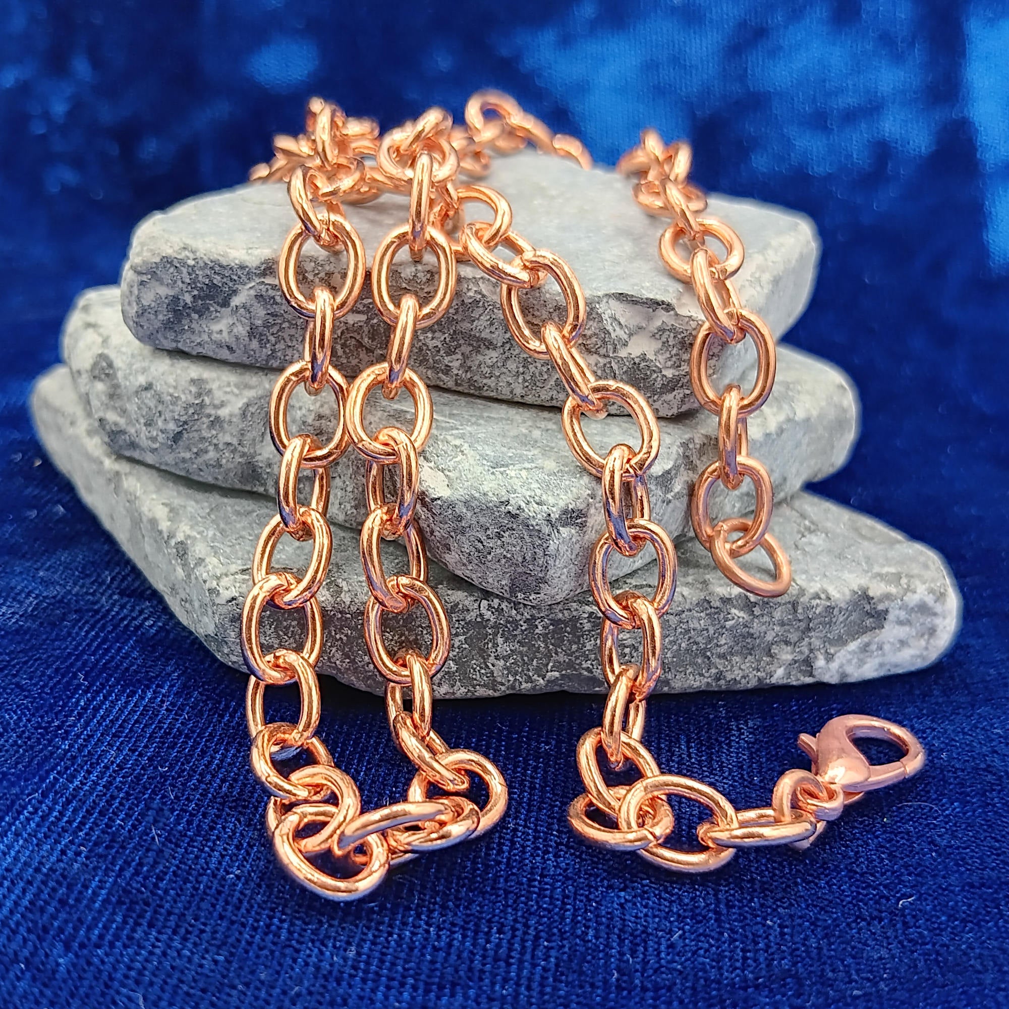 A heavy copper cable chain necklace draped over stacked gray stones on a textured blue fabric background.