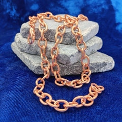 A copper chain link necklace draped over a stack of three gray stones on a textured blue fabric background.