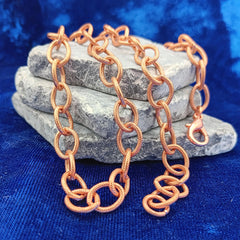 A copper chain link necklace with a lobster clasp, draped over a stack of gray stones on a textured blue fabric background.