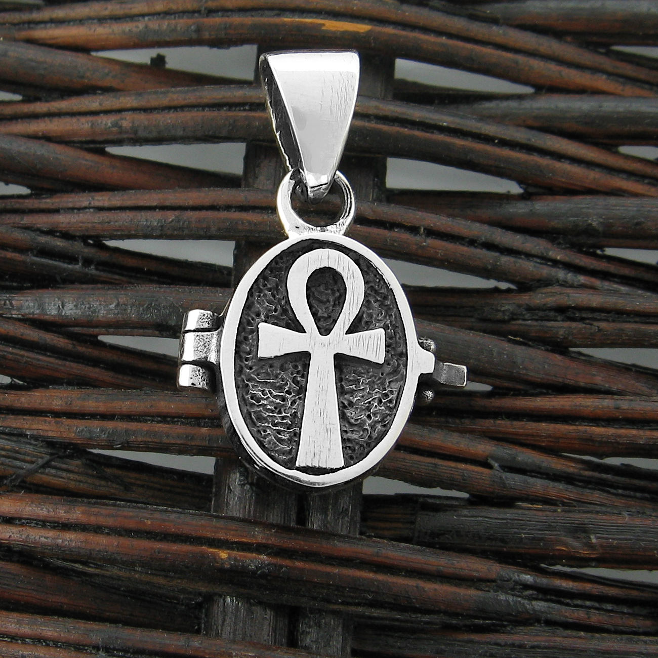 Front view of a small silver locket with a raised Ankh symbol resting on a dark woven basket.