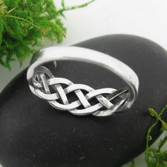 Back/inside view of a silver ring with a braided Celtic knot design resting on a flat black stone with green grass behind and a grayish white background.
