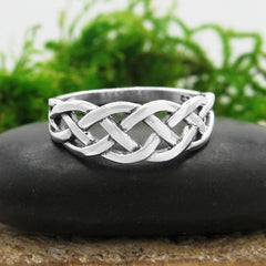 Front view of a silver ring with a braided Celtic knot design resting on a flat black stone with blurred green grass behind and a grayish white background.