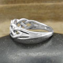Side view of a silver ring with a braided Celtic knot design resting on a flat black stone with blurred wood in the background