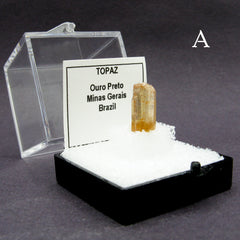 Side view of specimen of Brazilian 'honey' topaz gemstone labeled 'A' in plastic case on dark background.
