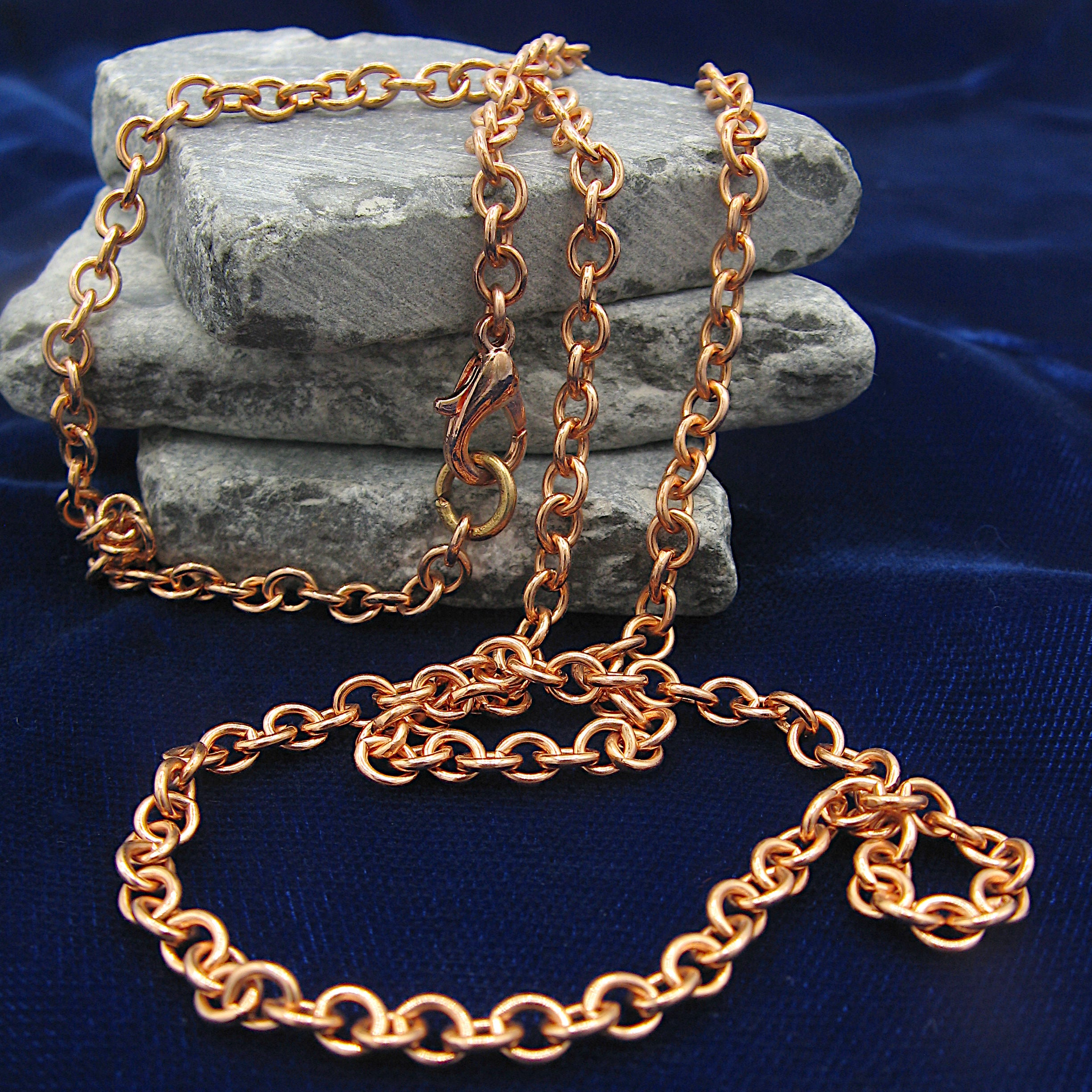 A copper cable chain with lobster clasp casually draped over  flat grey rocks with a dark blue background.