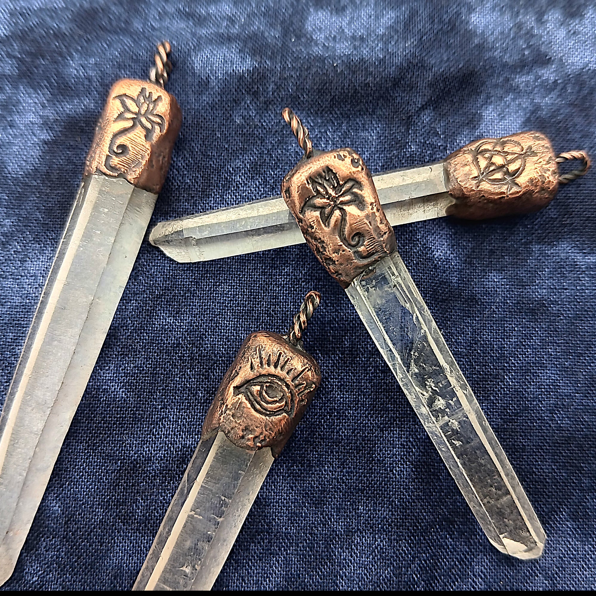 Four clear quartz crystal point pendants with ornate copper metal caps featuring symbolic engravings, arranged on a textured navy blue fabric background.