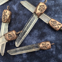 Four clear crystal point pendants with copper metal caps featuring engraved symbols, arranged on a dark blue textured fabric background.