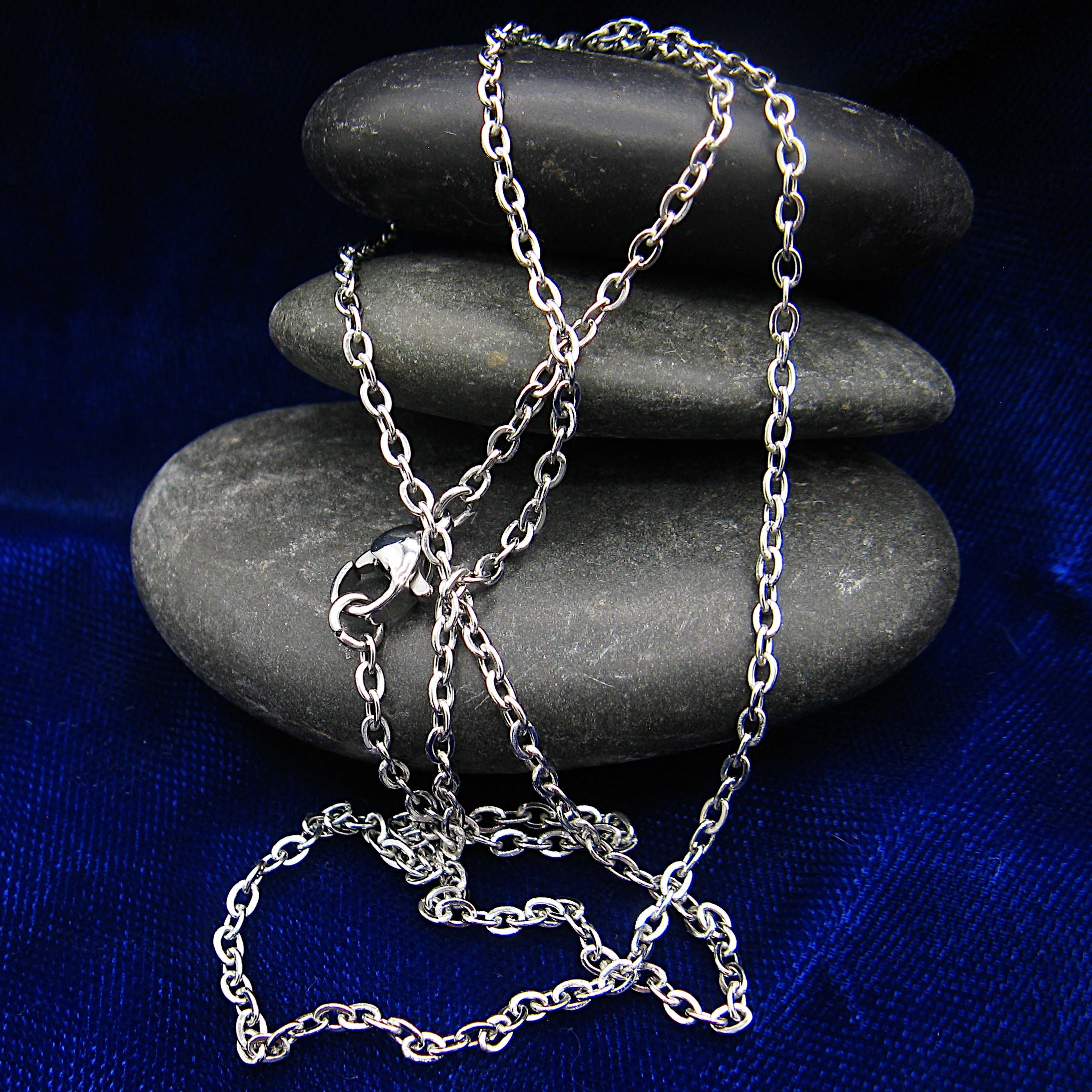 A silver stainless steel chain necklace with lobster clasp draped over a stack of smooth dark gray stones on a deep blue fabric background.