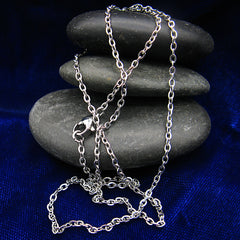 A silver stainless steel chain necklace with lobster clasp draped over a stack of smooth dark gray stones on a deep blue fabric background.
