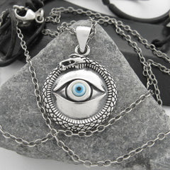 Front view of silver and blue evil eye pendant with snake border on a chain on a stone with black and white background.