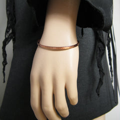 Close up view of a gently hammered narrow copper open-cuff bracelet worn by a mannequin wearing dark clothes against a gray background