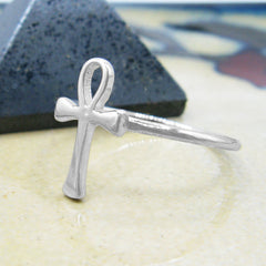 Angled side view of a silver ring in the shape of an Egyptian ankh symbol resting on a shiny tan surface with a floral pattern and a small dark pyramid behind it.