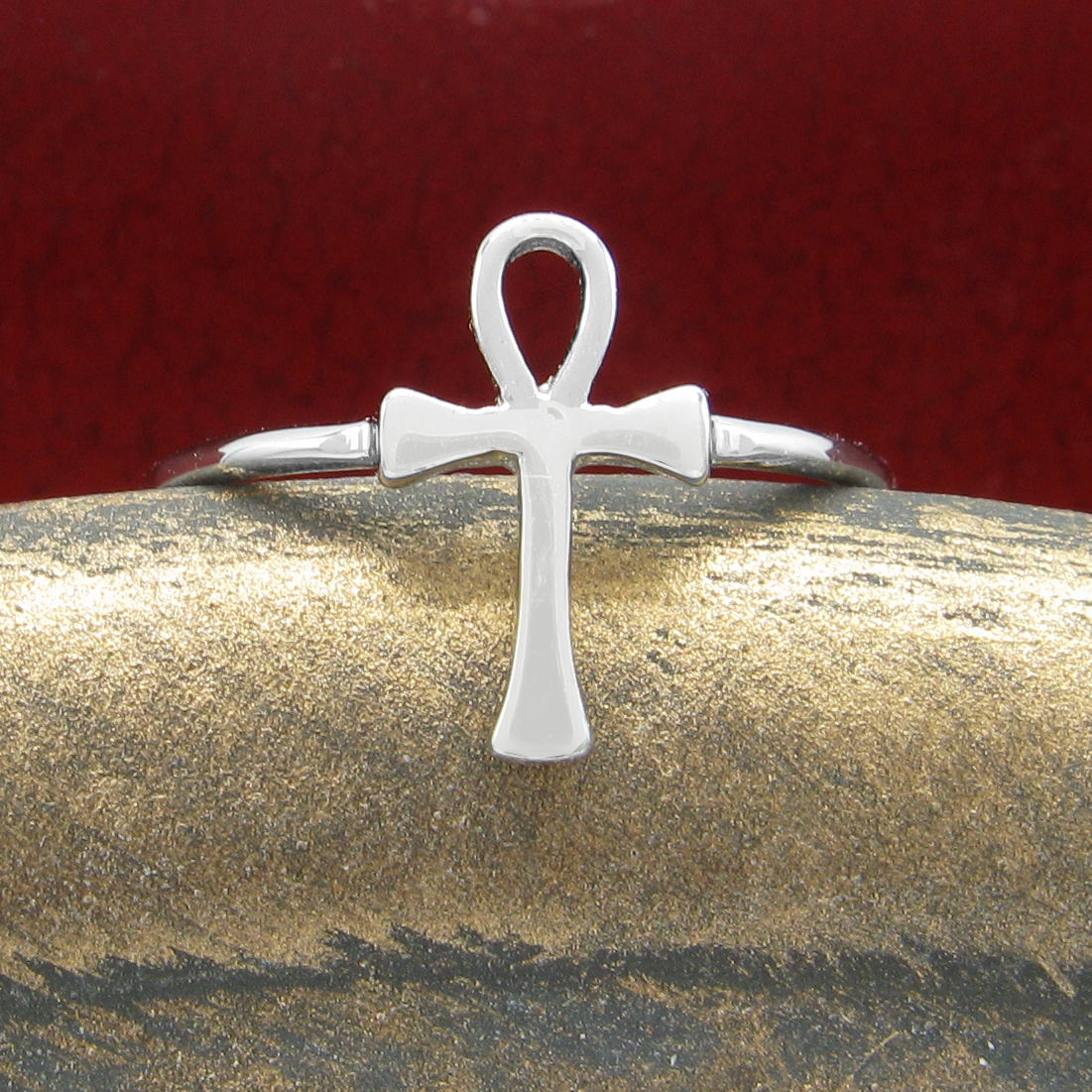 Front view of a silver ring in the shape of an Egyptian ankh symbol resting on a rounded textured golden ceramic prop with a dark red background.
