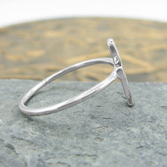 Side view of a silver ring in the shape of an Egyptian ankh symbol resting on a flat gray stone with a round golden prop in the background.