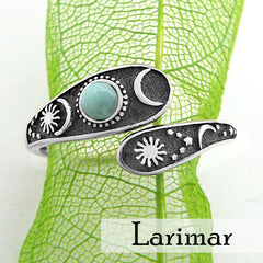 Front view of a wrap-around two-tiered oxidized silver ring with sun, moon, and stars design  and bluish-green cabochon on lacy green leaf with white background