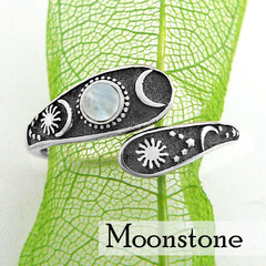 Front view of a wrap-around two-tiered oxidized silver ring with sun, moon, and stars design  and opalescent cabochon on lacy green leaf with white background