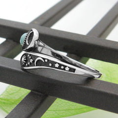 Side view of a wrap-around two-tiered oxidized silver ring with sun, moon, and stars design  and bluish-green cabochon resting on black metal frame with white background