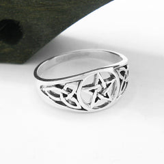 Angled view from slightly above of cut-out sterling silver Celtic style ring with pentagram and elongated triquetra knot resting on white background with part of dark wooden prop in one corner.