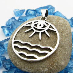 Back view of round cutout silver pendant with eye/sun/ocean design on smooth stone on random pieces of blue glass on white background