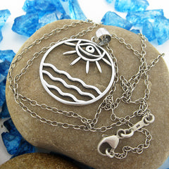 Front view of round cutout silver pendant with eye/sun/ocean design with chain on smooth stone on random pieces of blue glass on white background