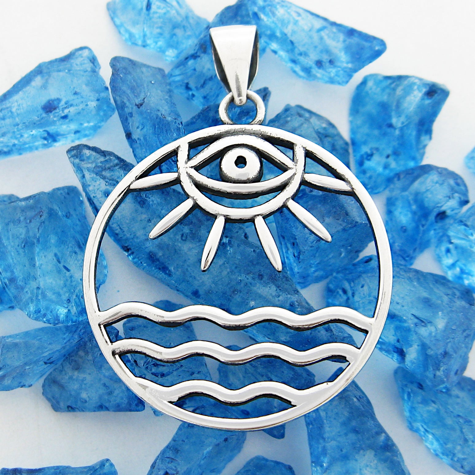 Front view of round cutout silver pendant with eye/sun/ocean design on random pieces of blue glass on white background