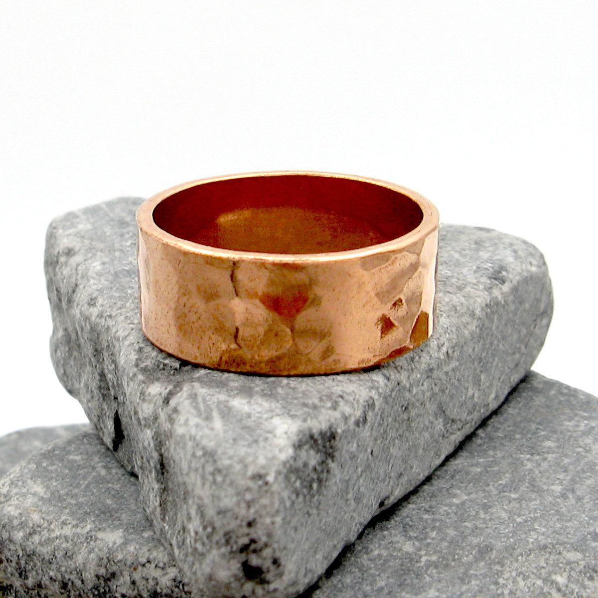 Gently hammered pure copper band ring resting on stacked grey stones with white background.