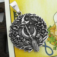 Front view of a round oxidized silver pendant with witch's broom and hat and crescent moon surrounded by flowers and stars resting on a yellow Tarot card with a wooden prop in the background