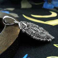 Side view of a round oxidized silver pendant with a textured design leaning against a wooden prop on a dark cloth background with blue and ivory pattern