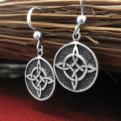 Angled side view of two round silver hook earrings with oxidized Witch's Knot design hanging upright from the bristles of an old fashioned broom lying horizontally on a glazed red ceramic surface.