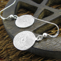 View of backs of two round silver hook earrings with hammered finish and 925 inscription lying flat on a round dark wooden lattice-like prop on top of the bristles of an old-fashioned broom.